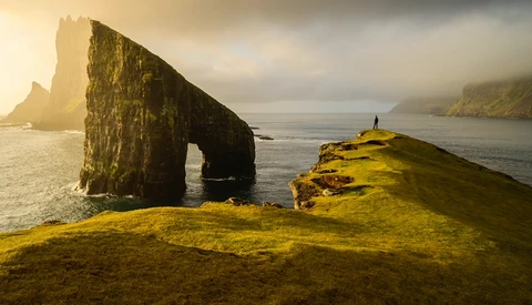 Moss-covered coastal cliff with natural rock arch framing calm sea and distant headlands.