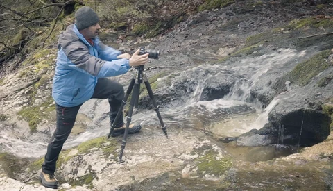 Photographer on tripod shooting a flowing stream in a moss-covered woodland setting.