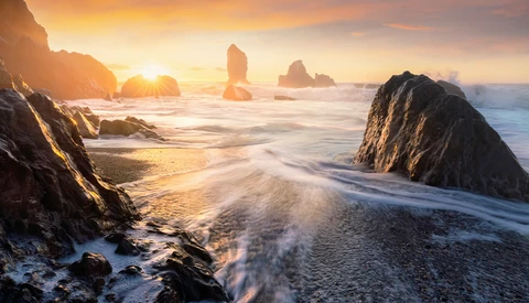 Rocky sea stacks silhouetted against golden sunrise over a misty ocean beach.