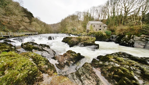 Moss-covered rocks frame a fast-flowing river with a stone cottage nestled among bare trees on the hillside.