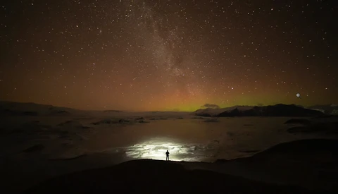 Silhouetted figure standing on moonlit beach beneath star-filled sky and aurora glow.