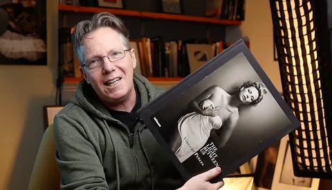 Photographer holding a black and white portrait book in a studio setting with bookshelves behind.