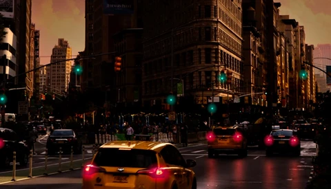 Yellow taxi cabs with illuminated taillights on a wet urban street at dusk, with traffic lights and tall buildings lining the road.