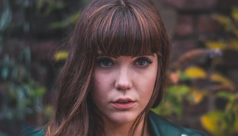 Woman with dark hair and bangs looking directly at camera against blurred foliage backdrop.