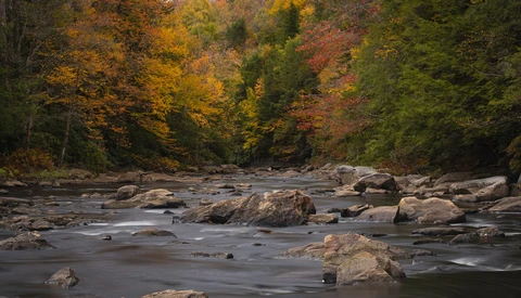 Rocky river with autumn foliage reflected in calm water and forest backdrop.
