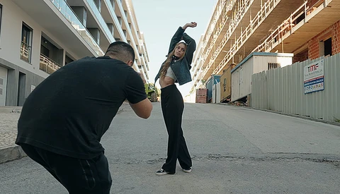 Photographer crouching with camera facing a model in black clothing posing on an urban street between buildings.