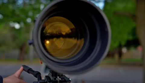 Close-up view through a rifle scope's golden-amber lens showing a blurred outdoor background with trees and buildings.