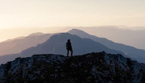 Silhouetted hiker standing on a rocky outcrop overlooking layered mountain ranges at sunset.