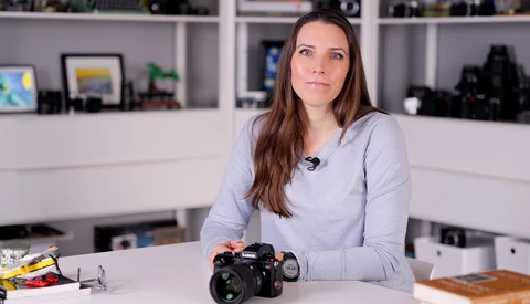 Woman presenter demonstrating a professional DSLR camera at a desk in a studio setting.