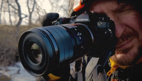 Photographer holding Olympus camera with telephoto lens against bare trees in outdoor setting.