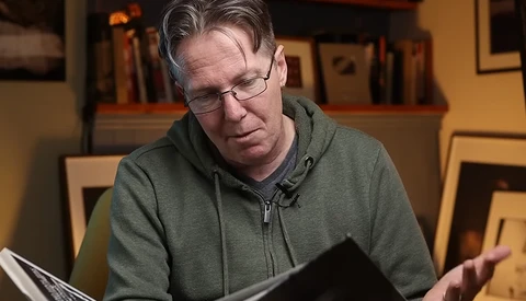 Man wearing glasses and olive green hoodie reviewing printed photographs at a table.