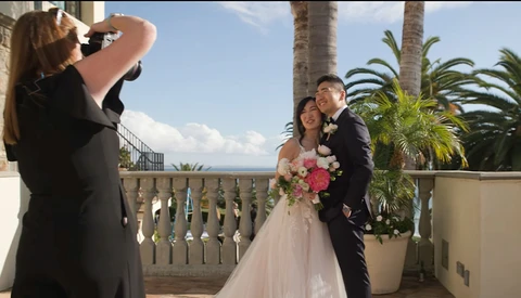 Wedding photographer capturing bride and groom posing on a terrace with palm trees and ocean view.