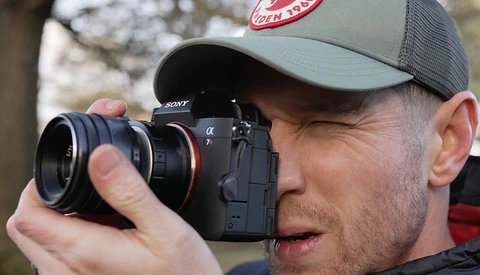 Photographer holding a Sony mirrorless camera with a telephoto lens, wearing a gray baseball cap.