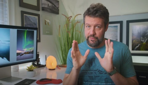 Man in teal shirt gesturing while discussing photography at desk with monitor displaying aurora image.