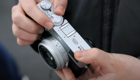 Hands holding a compact rangefinder camera with manual dials and a fixed lens.