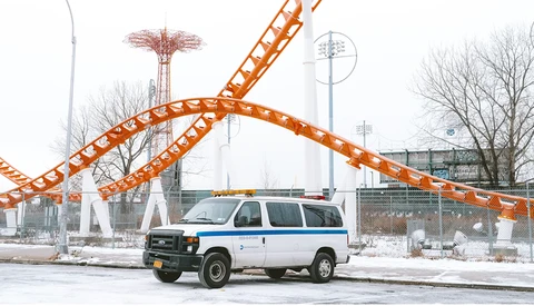White and blue van parked in front of an orange roller coaster structure in an amusement park setting.