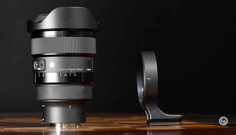 Wide-angle lens with lens hood and tripod collar mounted on wooden surface against black background.