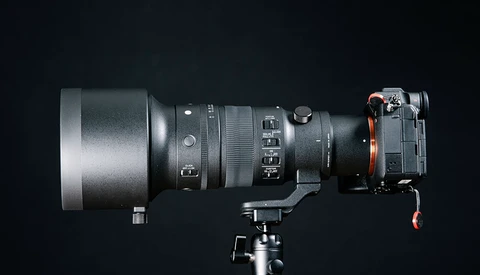 Professional camera lens mounted on tripod head with focus and zoom rings visible against dark background.