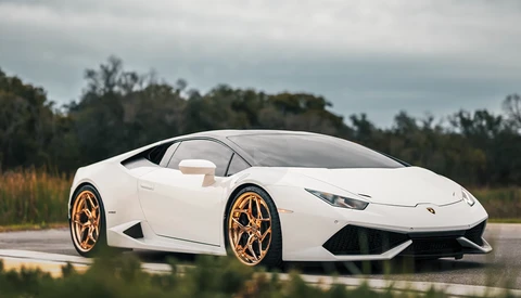 White sports car with gold wheels parked on wet pavement near trees.