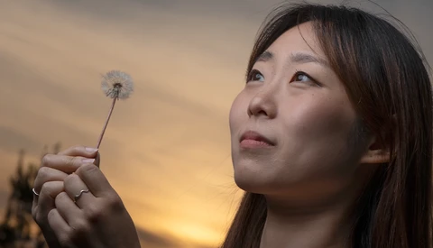 Woman holding a dandelion seedhead against a golden sunset sky.