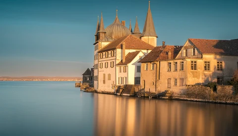 Yellow and cream-colored castle with multiple spires reflected in still blue water at golden hour.