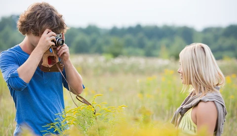 Young photographer with curly hair in blue shirt holding vintage camera up to face while photographing blonde woman in field.