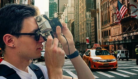 Photographer holding vintage film camera at street level, capturing urban cityscape with tall buildings and yellow taxi.