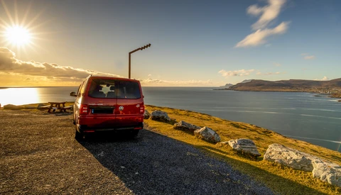 Red van parked on a coastal headland at sunset, overlooking a bay with mountains in the distance.