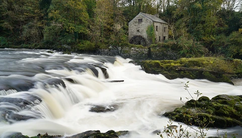Cascading waterfall with stone mill building surrounded by trees on riverbank.