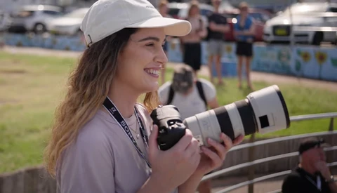 Female photographer holding a telephoto lens at an outdoor sports event.