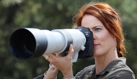 Photographer with reddish-brown hair holding a white telephoto lens to her eye while looking through the viewfinder.