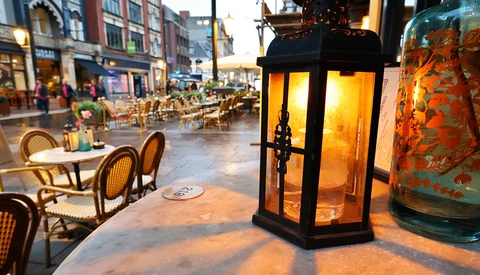 Illuminated black lantern on outdoor café table at dusk with blurred storefronts and diners in background.