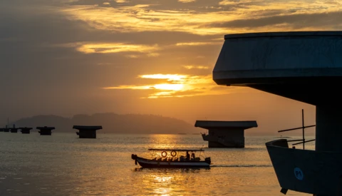 Silhouetted boats anchored in calm water at golden hour sunset with mountains in distant background.