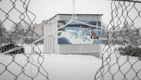 Winter mural viewed through chain-link fence showing snowman and figures in snowy landscape.