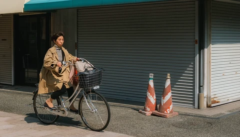 Woman on bicycle with basket stopped in front of shuttered storefront and traffic cones.