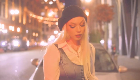 Woman with long blonde hair and dark beanie on urban street at night with warm city lights.