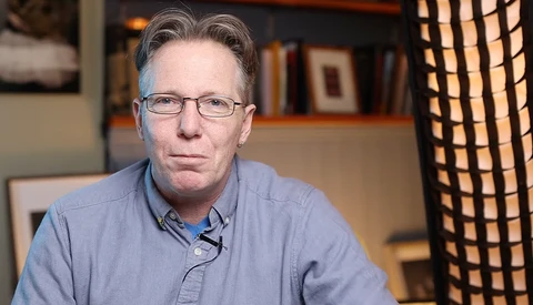 Man wearing glasses and blue button-up shirt seated indoors with bookshelves behind him.