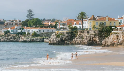 Swimmers in turquoise water below a clifftop coastal town with colorful buildings and sandy beach.