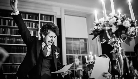 Groom in black tuxedo raising his fist triumphantly while holding papers in an elegant library setting.