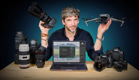 Male videographer and photographer surrounded by camera equipment and holding a drone and telephoto lens.