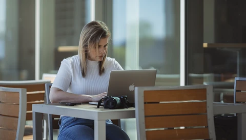 Woman working on laptop at outdoor table with city skyline in background.