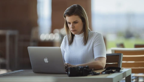 Woman working on MacBook at outdoor table with camera equipment nearby.