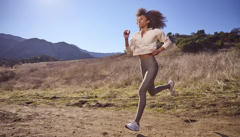 Woman jogging on an open trail through dry grassland with mountains in the distance under clear blue sky.
