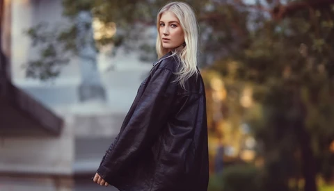 Woman with long platinum blonde hair wearing a dark navy jacket, photographed outdoors in soft golden-hour light.