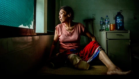 Woman in pink shirt sitting on floor in modest room, gazing toward window with natural light.