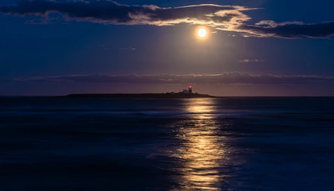 Full moon illuminating a golden path across calm ocean water toward a distant lighthouse at dusk.