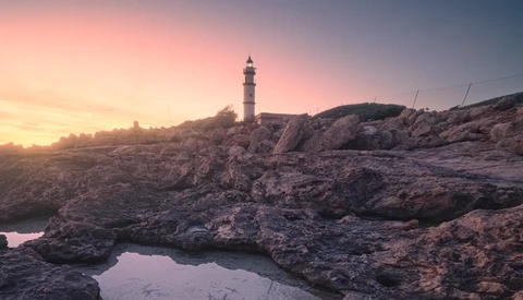 Lighthouse on rocky coastline at golden hour sunrise with tide pools in foreground.