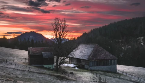 Two wooden chalets with snow-covered roofs sit in a winter valley beneath a dramatic pink and red sunset sky.