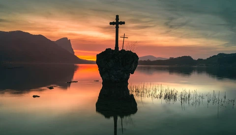 Silhouetted crosses on a rocky outcrop in still water at sunrise, with mountains and forested shorelines in the background.