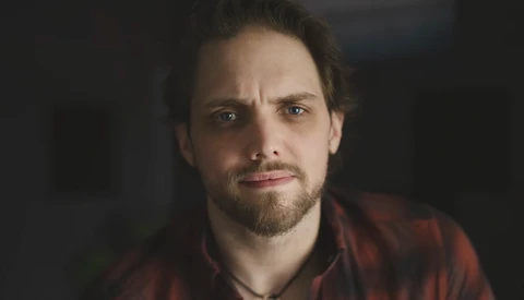 Man with dark hair and beard wearing a red plaid shirt, looking directly at camera with a serious expression.